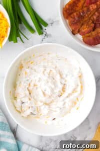 Creamy Loaded Baked Potato Dip Recipe for Parties 11 A white bowl filled with a sour cream based dip, with bowls of cooked bacon and cheese on the side.