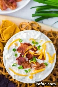 Creamy Loaded Baked Potato Dip Recipe for Parties 12 An overhead shot of a bowl of cream dip garnished with green onion, shredded cheese, and bacon. Surrounded by corn chips and pretzels.