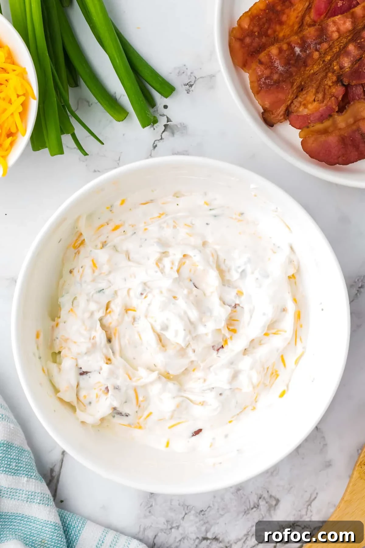 Creamy Loaded Baked Potato Dip Recipe for Parties 5 A white bowl filled with a sour cream based dip, with bowls of cooked bacon and cheese on the side.