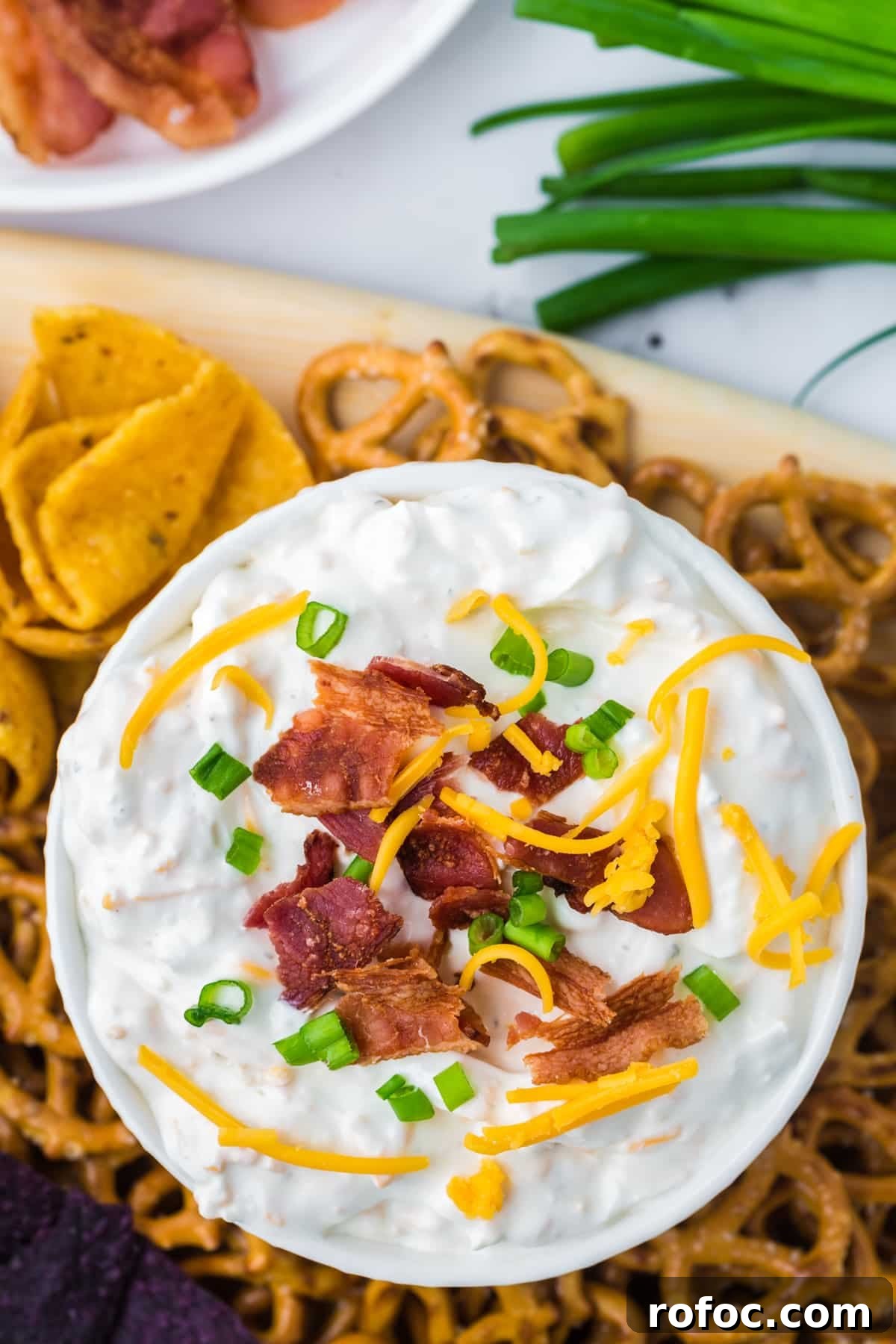 Creamy Loaded Baked Potato Dip Recipe for Parties 6 An overhead shot of a bowl of cream dip garnished with green onion, shredded cheese, and bacon. Surrounded by corn chips and pretzels.