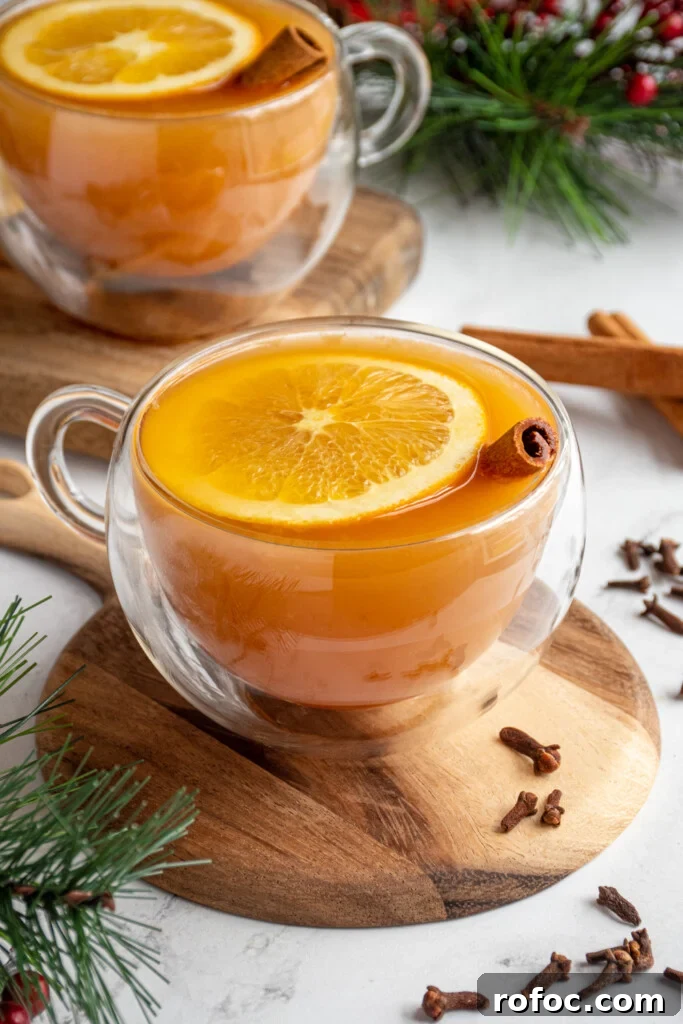 Slow-Cooker Christmas Wassail Recipe for Festive Parties 3 A clear glass mug filled with warm wassail garnished with an orange slice and cinnamon stick with cloves scattered about the table it is on.