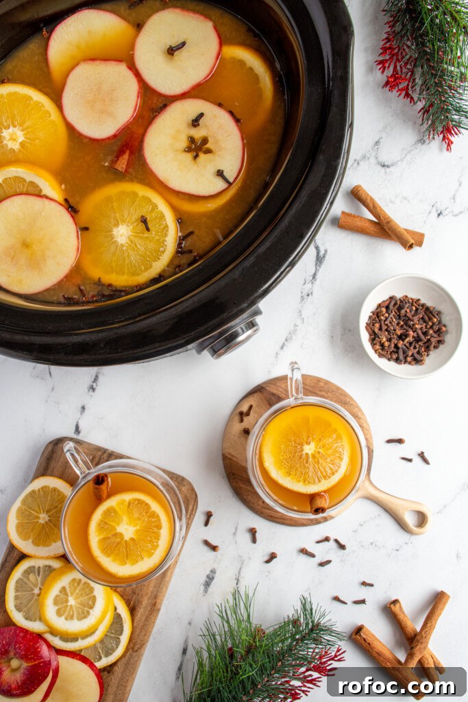 Slow-Cooker Christmas Wassail Recipe for Festive Parties 4 Overhead view of wassail in a crockpot next to full glasses of Christmas wassail in clear mugs topped with orange slices and cinnamon sticks.