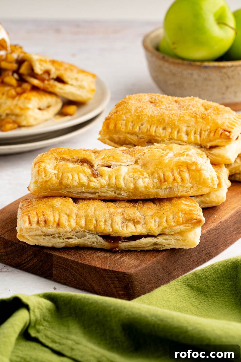 Homemade Apple Hand Pies: Flaky Mini Pastry Recipe 7 A bunch of apple hand pies stacked on top of a wood cutting board with a bowl of granny smith apples on the table behind them.