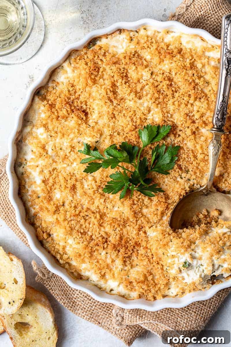 Creamy Baked Crab Dip with Garlic and Parmesan 2 baking dish with hot crab dip and serving spoon next to baguette crostini and glass of wine.