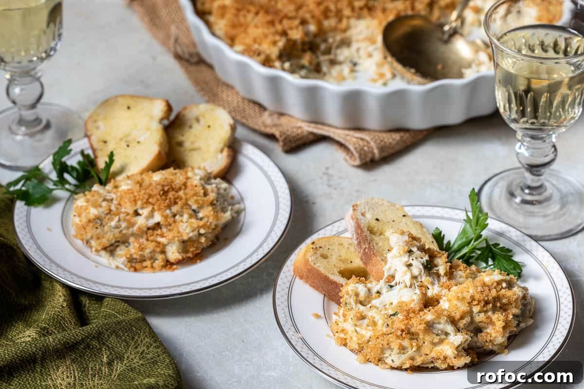Creamy Baked Crab Dip with Garlic and Parmesan 10 two small plates with crab dip and crostini toasts next to glasses of wine, in front of baking dish with crab dip.