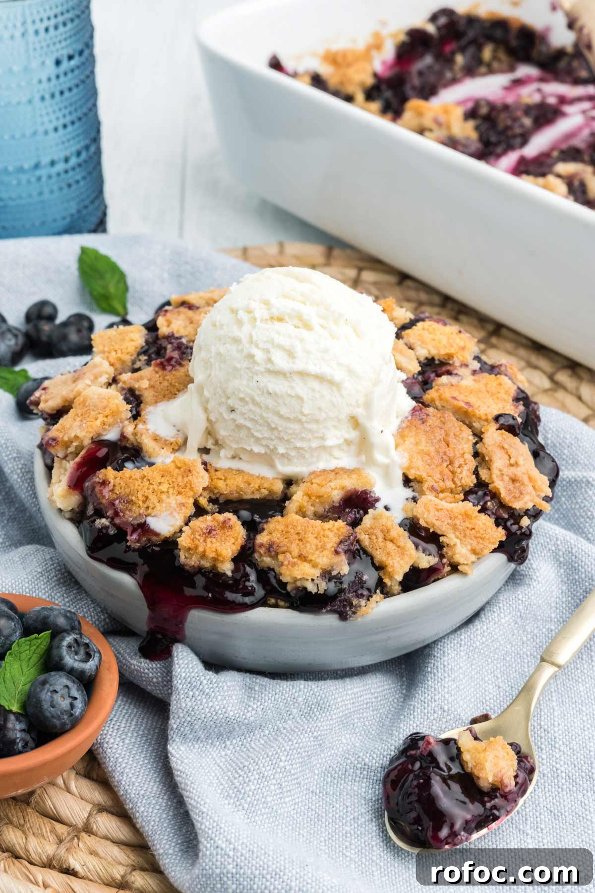 Blueberry Cake Mix Cobbler: Quick, Moist Dessert Recipe 8 Round dish of blueberry dump cake on a blue napkin with a scoop of vanilla ice cream on top.