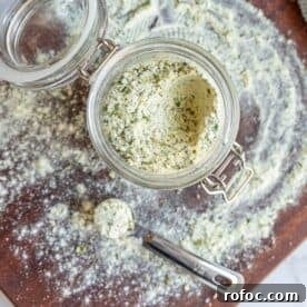 Homemade Ranch Seasoning Mix Recipe for Dip, Dressings & Marinades 7 Glass jar of herb-infused salt on a wooden surface with spilled salt and a measuring spoon.