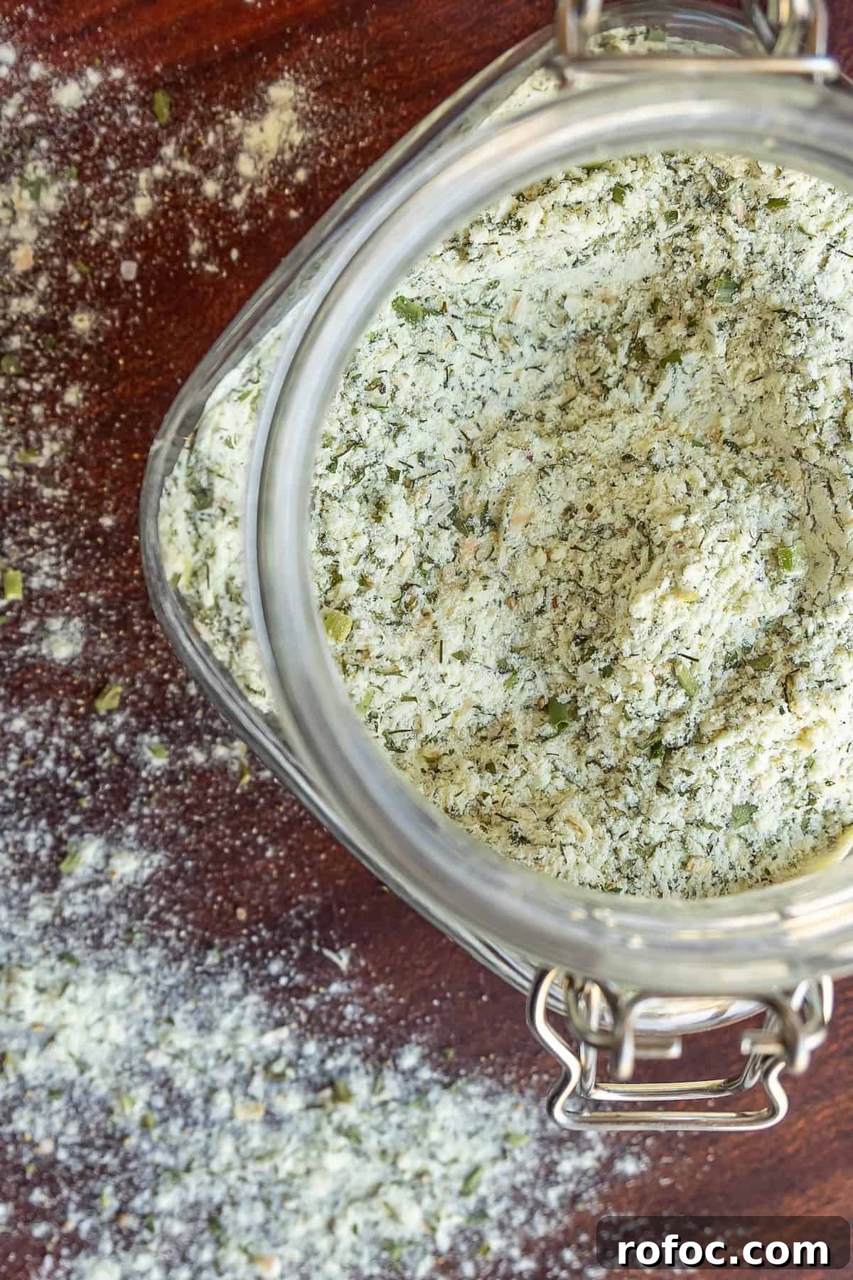 Homemade Ranch Seasoning Mix Recipe for Dip, Dressings & Marinades 3 Open glass jar filled with a dry herb and seasoning blend, with some spilled on a wooden surface.