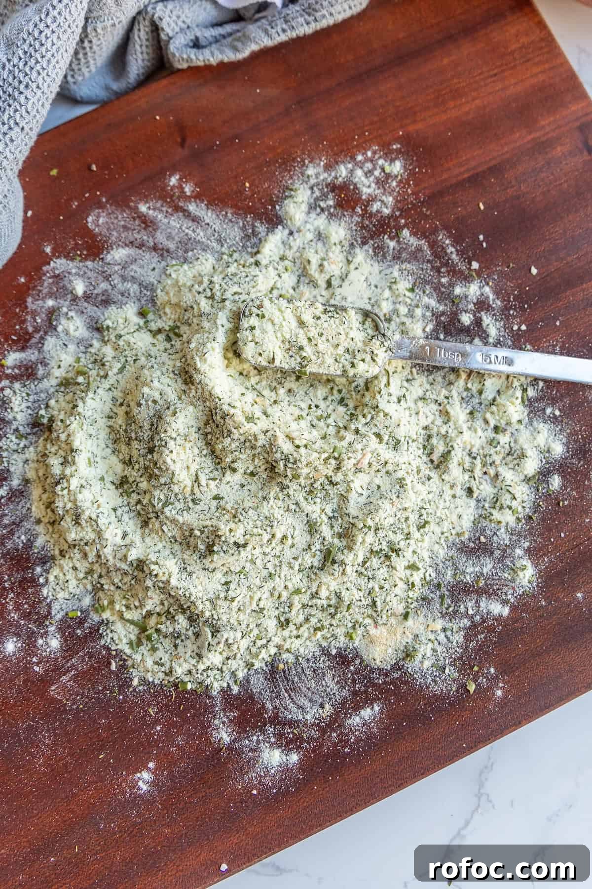 Homemade Ranch Seasoning Mix Recipe for Dip, Dressings & Marinades 6 A mix of herbs and flour with a teaspoon on a wooden surface. A gray cloth is partially visible in the upper left corner.