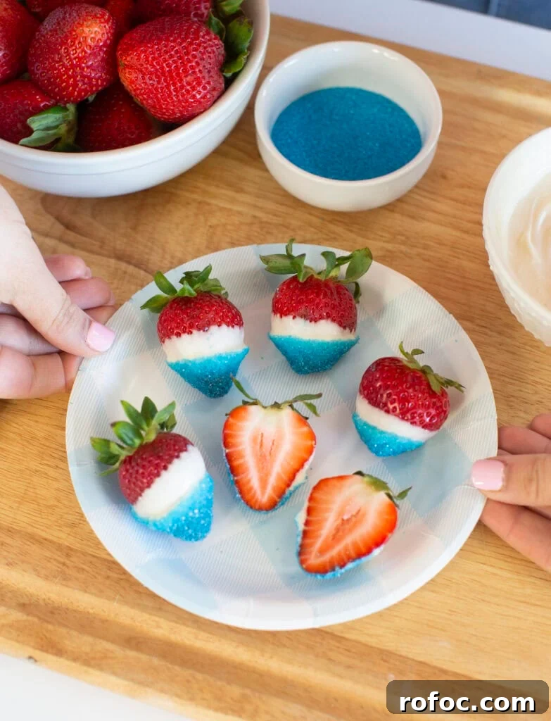 Patriotic Dipped Strawberries for 4th of July Celebrations 2 A plate of strawberries dipped in white chocolate and blue sugar sits on a wooden surface. Two hands hold the plate. A bowl of plain strawberries, a bowl of blue sugar, and a bowl of white chocolate are nearby.