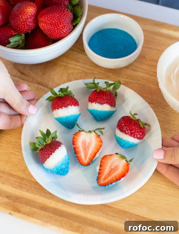 Patriotic Dipped Strawberries for 4th of July Celebrations 10 A plate of strawberries dipped in white chocolate and blue sugar sits on a wooden surface. Two hands hold the plate. A bowl of plain strawberries, a bowl of blue sugar, and a bowl of white chocolate are nearby.