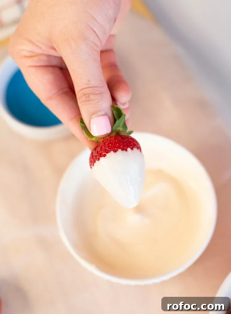 Patriotic Dipped Strawberries for 4th of July Celebrations 4 A hand with pink nail polish holds a strawberry partially dipped in white chocolate above a white bowl filled with melted white chocolate.