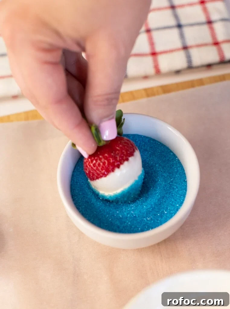 Patriotic Dipped Strawberries for 4th of July Celebrations 5 A hand dips a white chocolate-covered strawberry into a bowl of blue sugar, creating a red, white, and blue treat. A striped cloth is visible in the background.