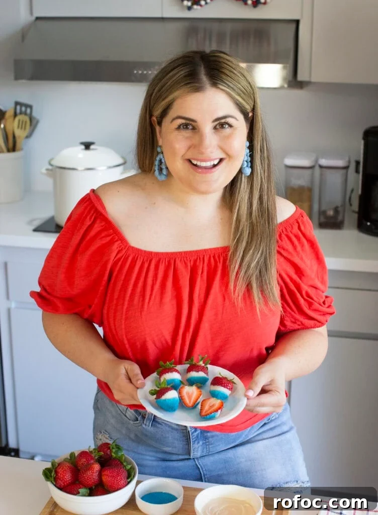 Patriotic Dipped Strawberries for 4th of July Celebrations 6 A woman in a red off-the-shoulder top holds a plate of decorated strawberries in a modern kitchen. Bowls of strawberries, blue sprinkles, and white chocolate are on the counter in front of her.