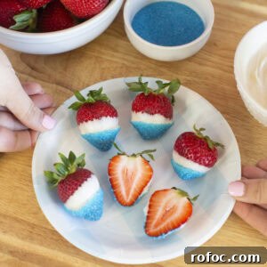 Patriotic Dipped Strawberries for 4th of July Celebrations 7 A plate of strawberries dipped in white chocolate and blue sugar sits on a wooden surface. Two hands hold the plate. A bowl of plain strawberries, a bowl of blue sugar, and a bowl of white chocolate are nearby.