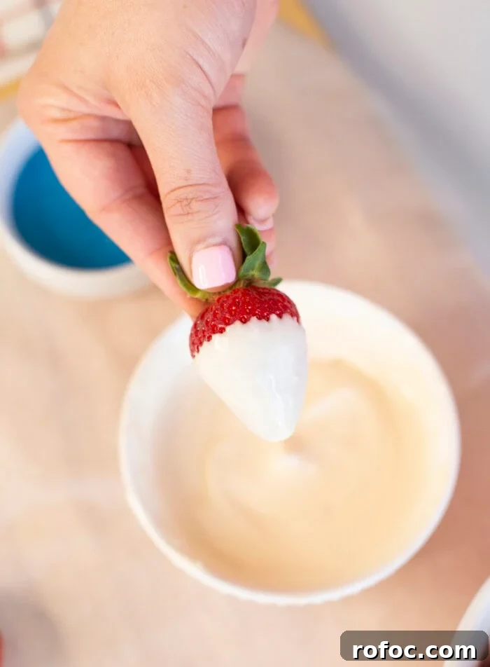 Patriotic Dipped Strawberries for 4th of July Celebrations 8 A hand with pink nail polish holds a strawberry partially dipped in white chocolate above a white bowl filled with melted white chocolate.