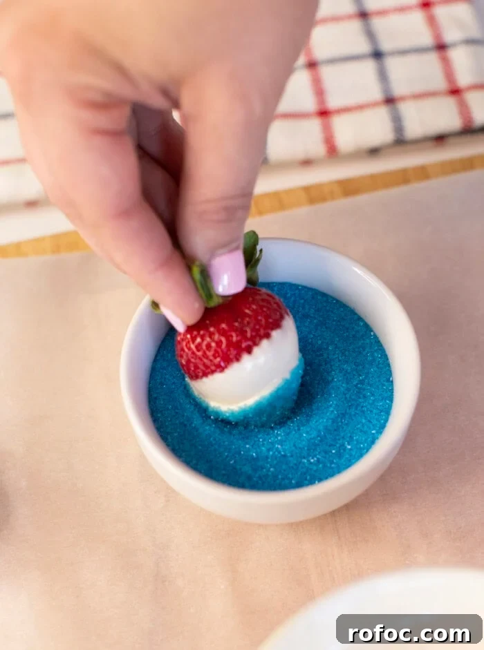 Patriotic Dipped Strawberries for 4th of July Celebrations 9 A hand dips a white chocolate-covered strawberry into a bowl of blue sugar, creating a red, white, and blue treat. A striped cloth is visible in the background.