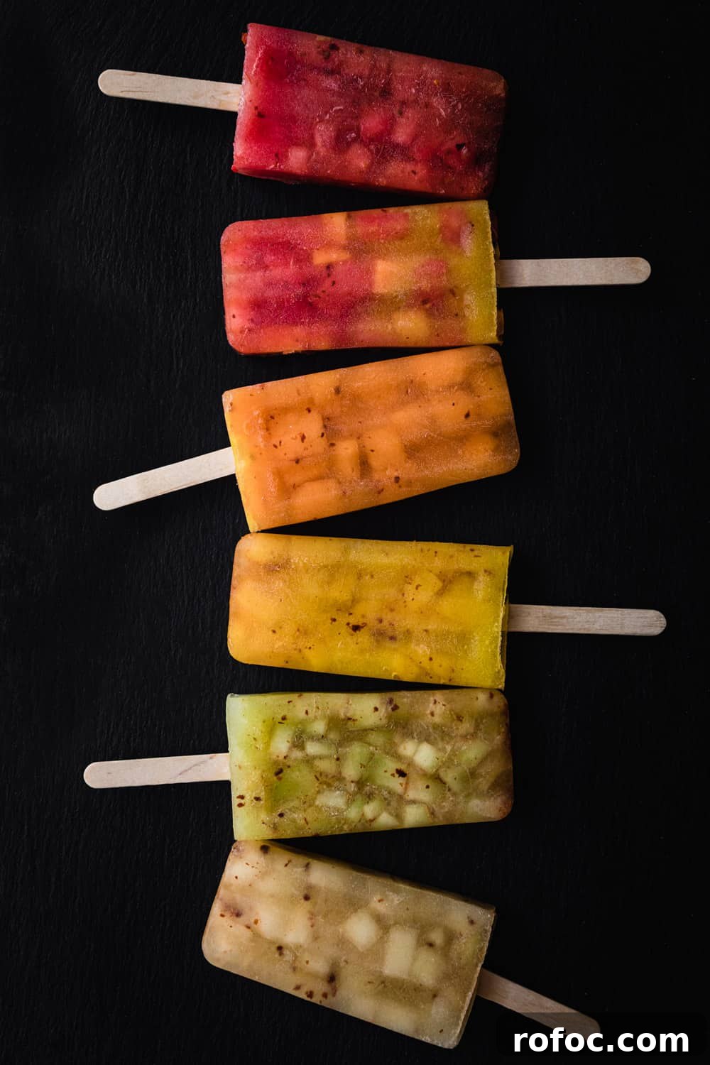 Spiced Watermelon Popsicles: Vibrant, Refreshing Summer Treat 7 5 melon popsicles in red, orange, yellow, green white and mixed colors lined up on a black background; overhead shot