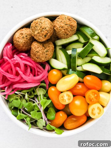 A large white bowl filled will cucumbers, tomatoes, greens, pink pickled red onions, and falafel.