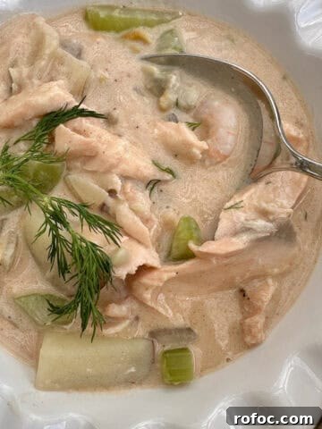 A bowl full of seafood chowder with a sprig of fresh dill and a spoon.
