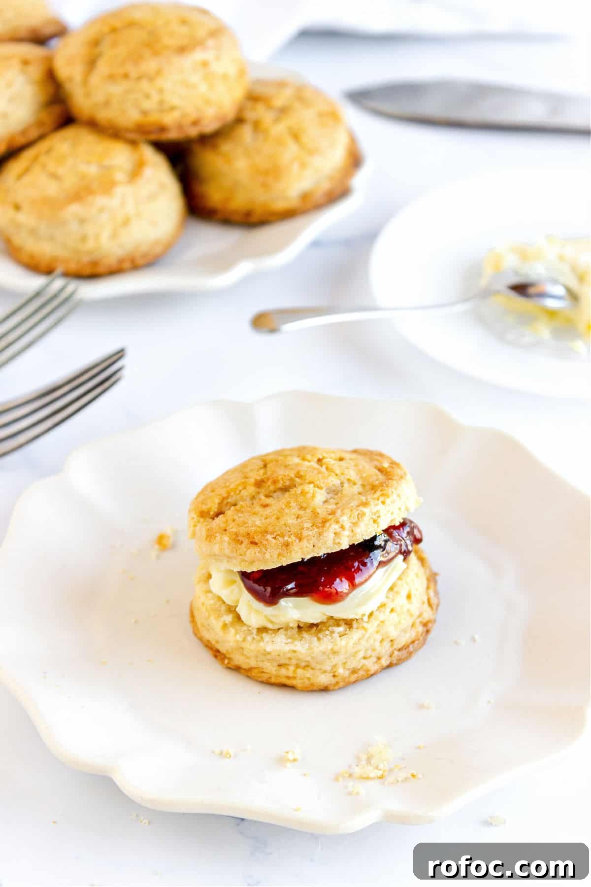 Mini Cream Scones Recipe — Simple Homemade Bite-Sized Scones 2 Butter and jam filled scone on a plate. White background with more scones and cutlery.
