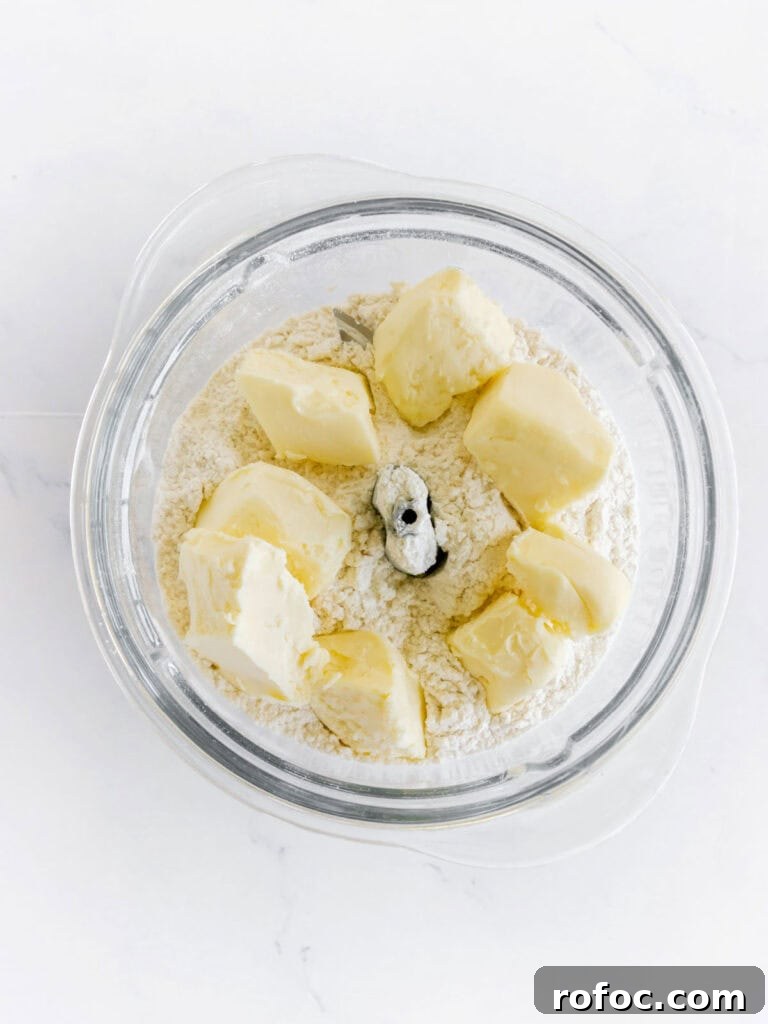 Mini Cream Scones Recipe — Simple Homemade Bite-Sized Scones 5 Butter pieces and dry ingredients in a food processor bowl on white marble.