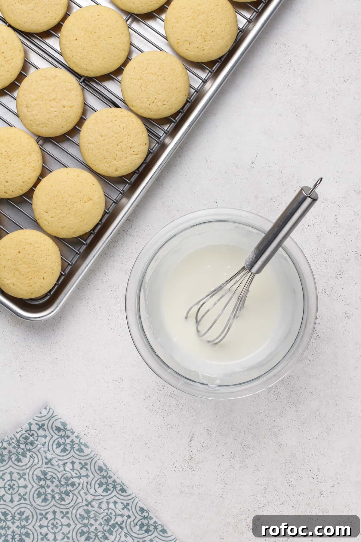 Lemon glaze in a glass bowl next to a cooling rack of cookies.