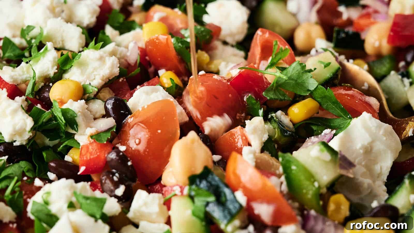 35 Gameday Recipes That Always Crowd-Pleasers Love 17 Close-up of a mixed salad with tomatoes, feta cheese, black beans, chickpeas, corn, cucumber, red onion, and parsley, being drizzled with dressing.