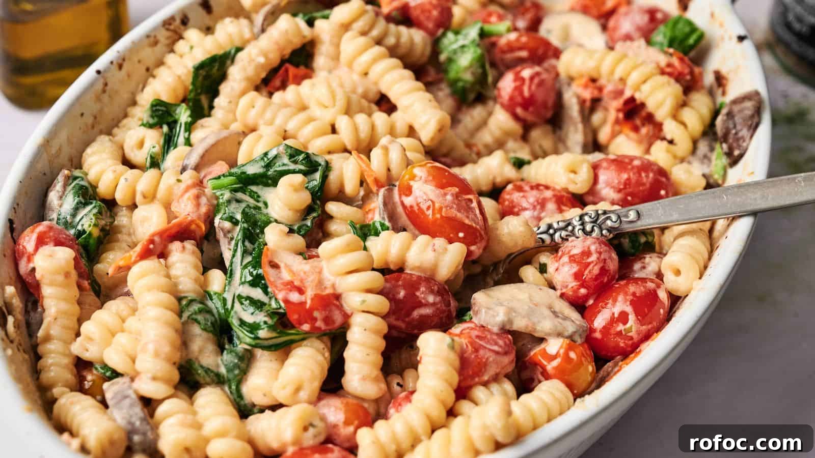35 Gameday Recipes That Always Crowd-Pleasers Love 31 A white dish filled with cooked fusilli pasta, cherry tomatoes, spinach, and mushrooms in a creamy sauce, with a silver fork resting inside.