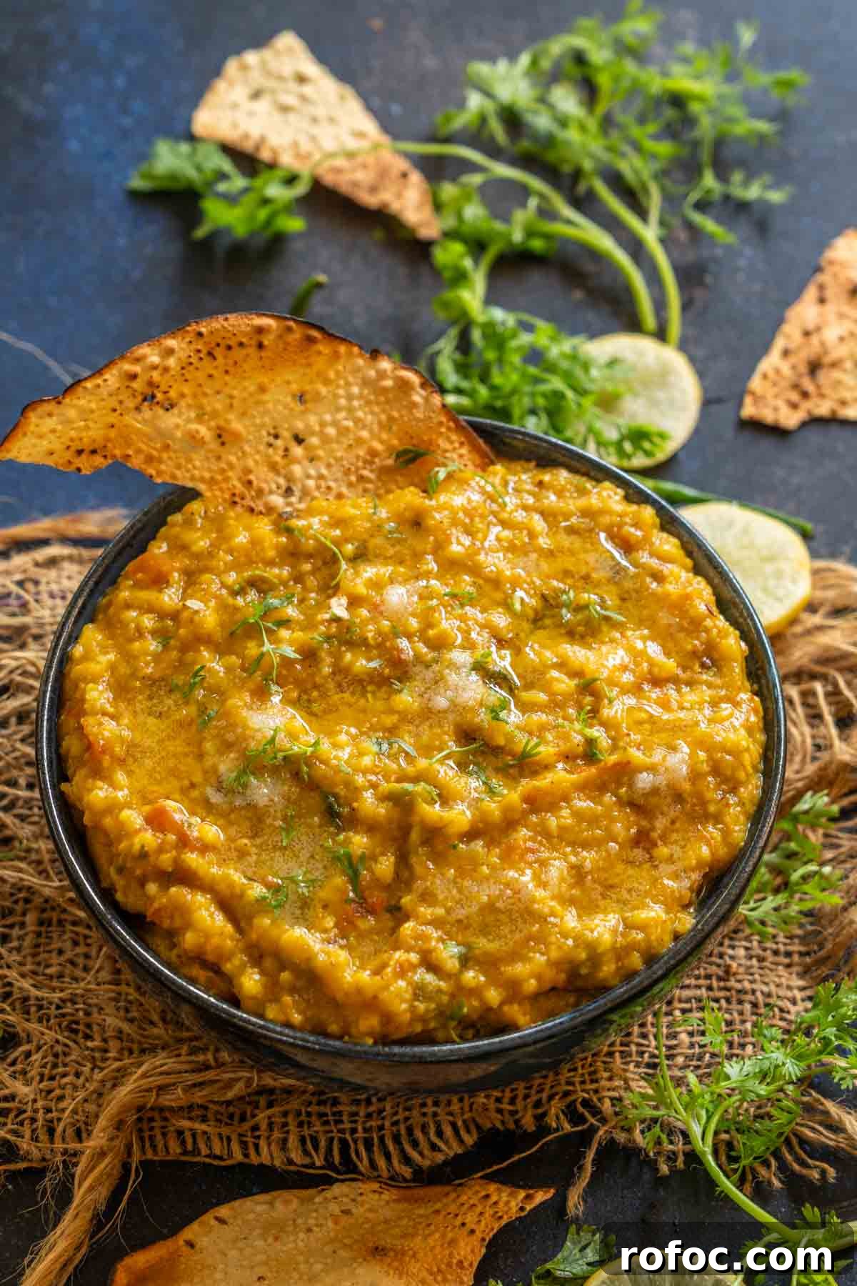 Jowar Khichdi Recipe: How to Cook Nutritious Sorghum Khichdi 2 A bowl of jowar khichdi garnished with fresh herbs, served alongside crispy papad and slices of lemon, chopped cilantro, and broken papad are scattered on a dark surface in the background.