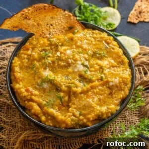 Jowar Khichdi Recipe: How to Cook Nutritious Sorghum Khichdi 8 A bowl of jowar khichdi garnished with fresh herbs, served alongside crispy papad and slices of lemon, chopped cilantro, and broken papad are scattered on a dark surface in the background.