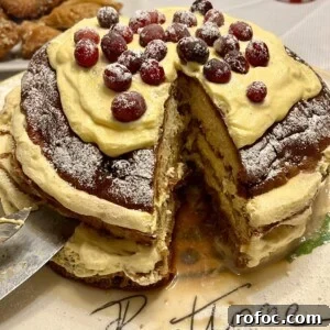 Panettone Tiramisu: Festive Holiday Dessert Recipe 3 A round cake with creamy layers, topped with powdered sugar and cranberries, sits on a plate with the word Panettone written on it. A slice is cut out, revealing the moist interior. In the background, there are pastries and a striped candle.