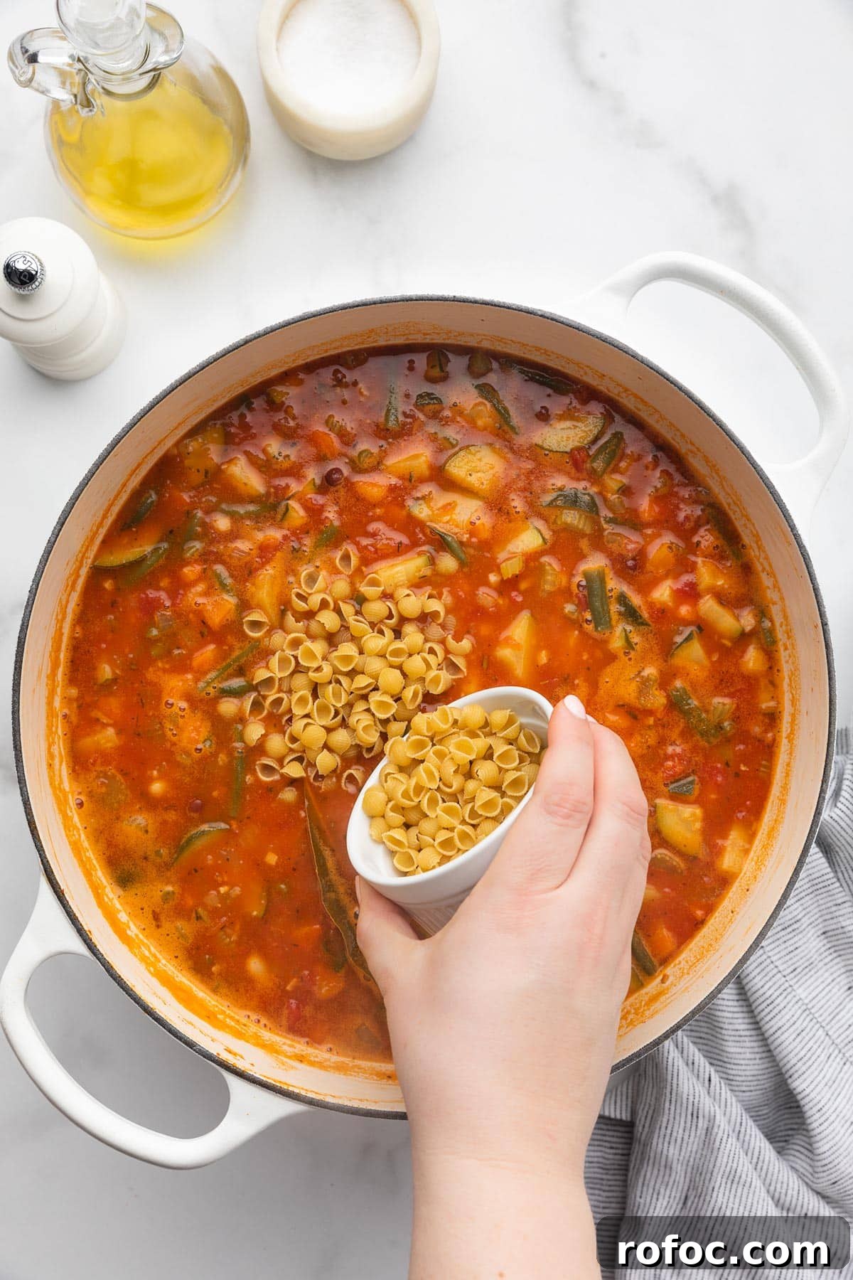 Classic Italian Minestrone Soup Recipe for Cozy Weeknight Dinners 13 pouring dry mini shell pasta into a pot of minestrone soup from a small white ramekin