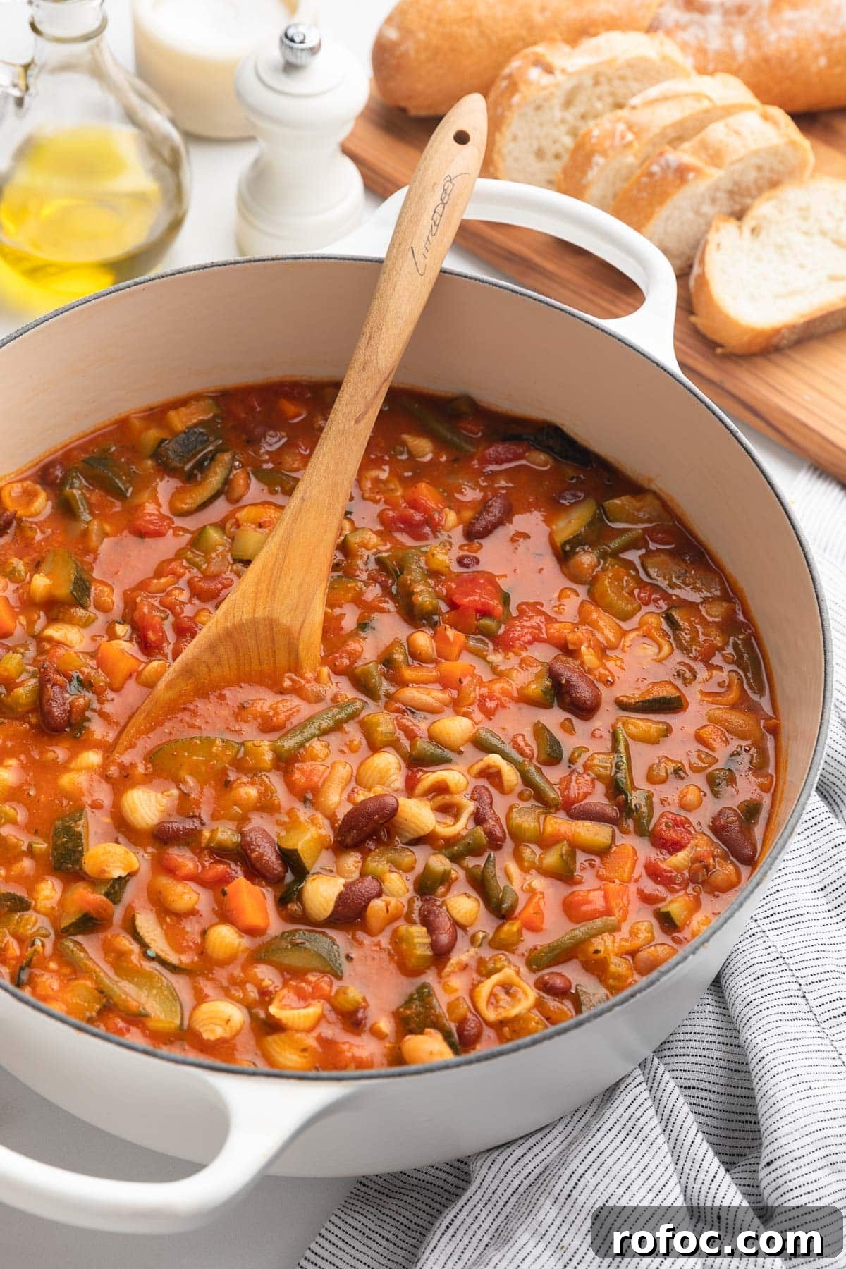 Classic Italian Minestrone Soup Recipe for Cozy Weeknight Dinners 17 minestrone soup in a white enameled cast iron Dutch oven with a wooden spoon next to a cruet of olive oil and a wood board of sliced Italian bread
