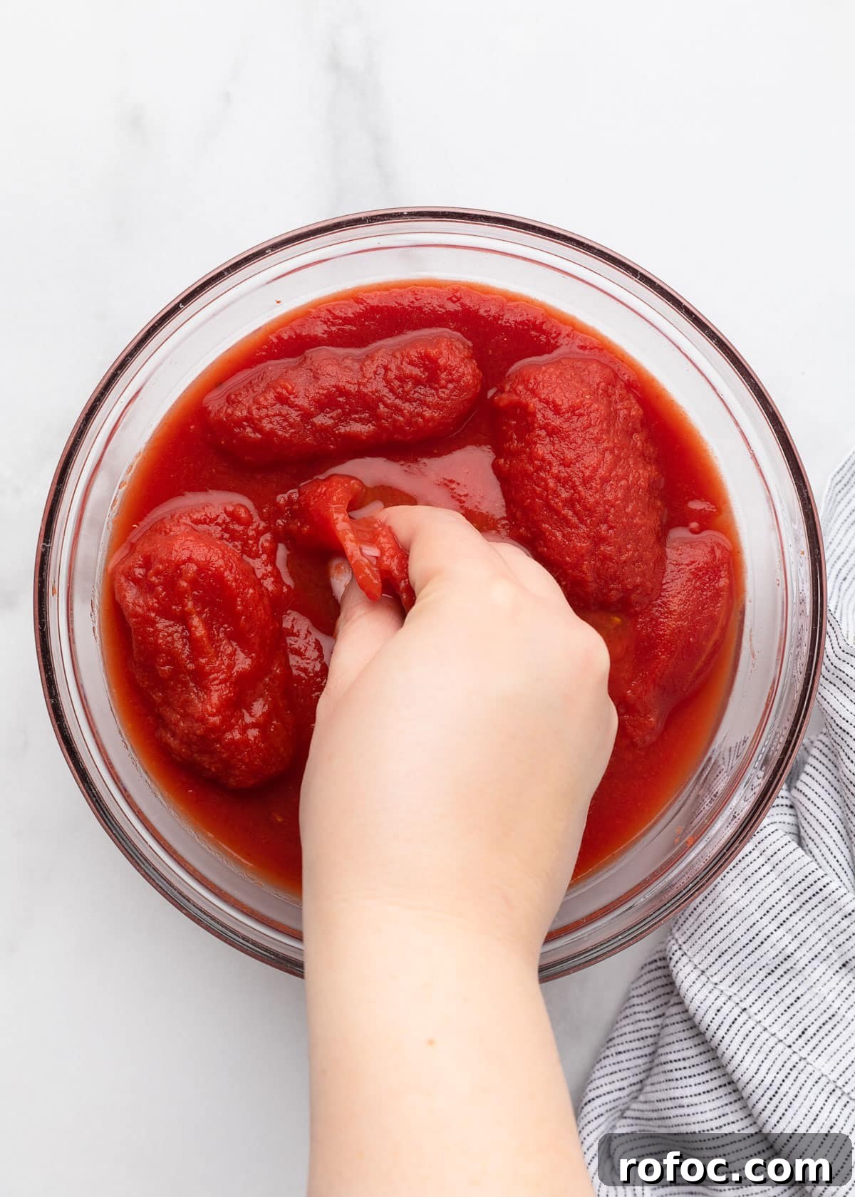 Classic Italian Minestrone Soup Recipe for Cozy Weeknight Dinners 4 hand crushing canned plum tomatoes in a glass bowl on a white marble surface