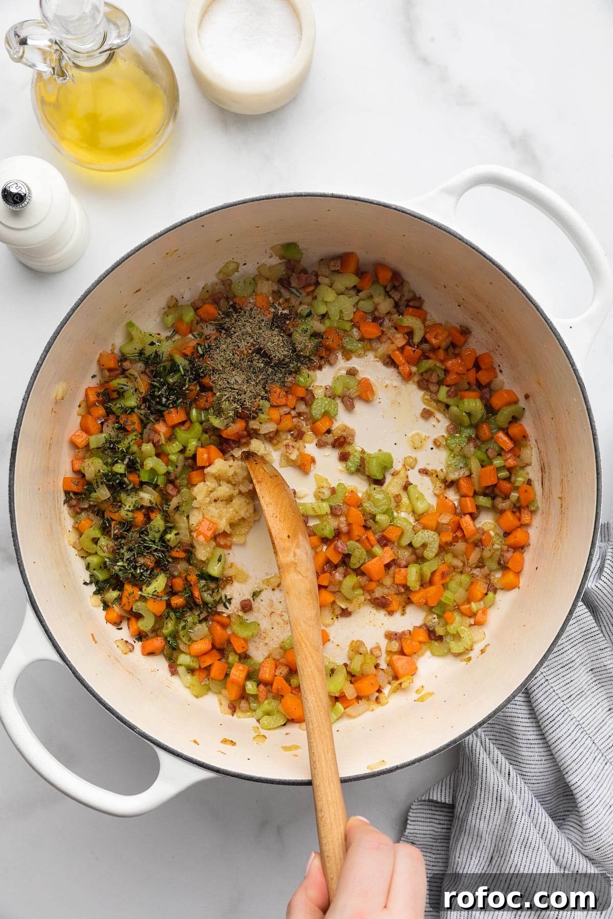 Classic Italian Minestrone Soup Recipe for Cozy Weeknight Dinners 8 minced garlic and herbs being stirred into cooked mirepoix in a white enameled cast iron Dutch oven with a wooden spoon