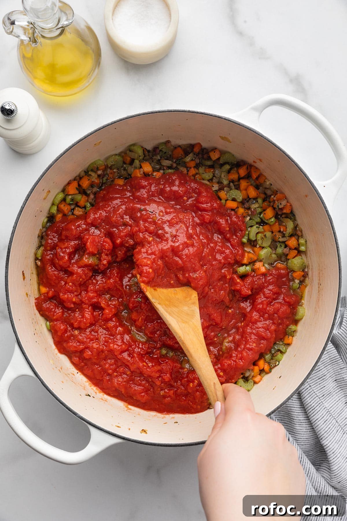 Classic Italian Minestrone Soup Recipe for Cozy Weeknight Dinners 9 crushed tomatoes being stirred into cooked mirepoix in a white enameled cast iron Dutch oven with a wooden spoon