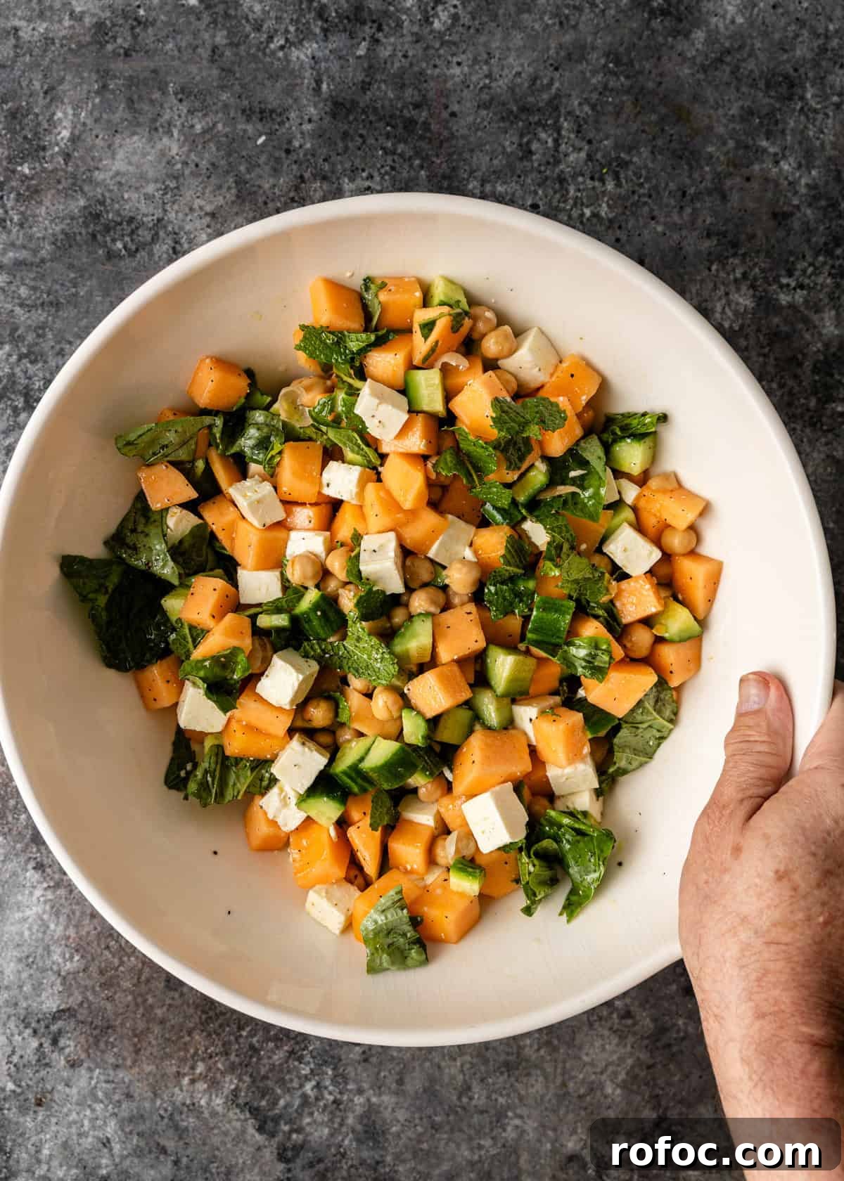 overhead: a hand holding a large white bowl of cantalope salad