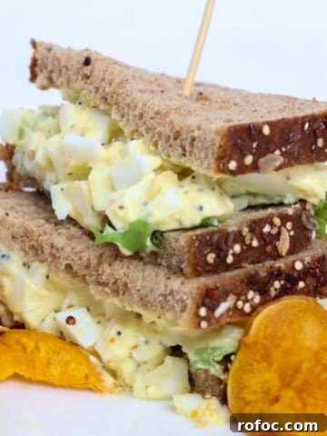 Two halves of an egg salad sandwich stacked, on a plate with sweet potato chips.