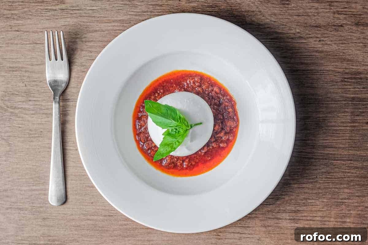Spicy Nduja with Creamy Burrata: Fiery Italian Cheese Pairing 2 A plate of nduja sausage in liquid form, topped with a ball of burrata cheese and garnished with a small sprig of basil. A fork sits to the left.