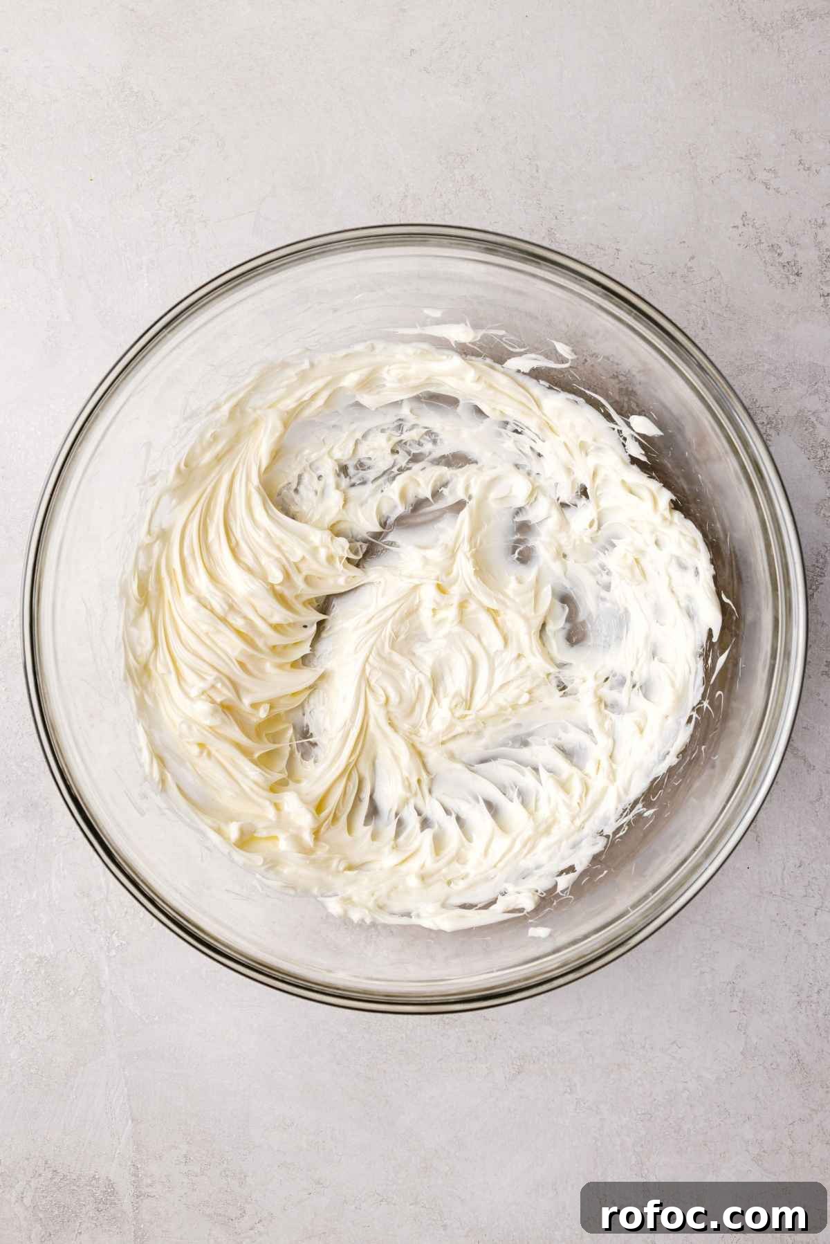 Unsalted butter whisked until fluffy in a large glass mixing bowl on a white and grey textured background.
