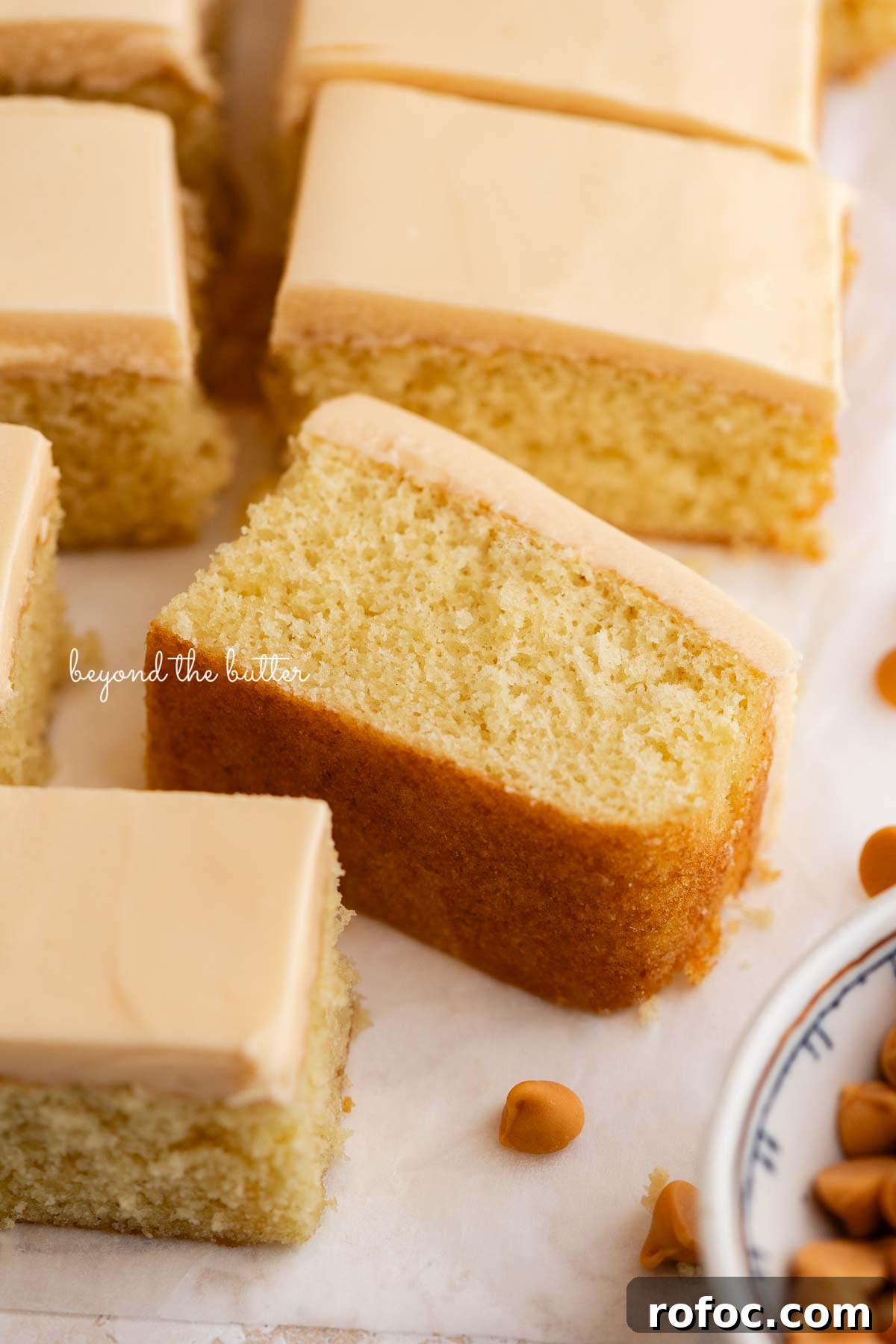 Slices of butterscotch krimpets placed on a parchment paper lined background with one slice on its side with a small decorative ceramic bowl of butterscotch chips placed next to the cake.