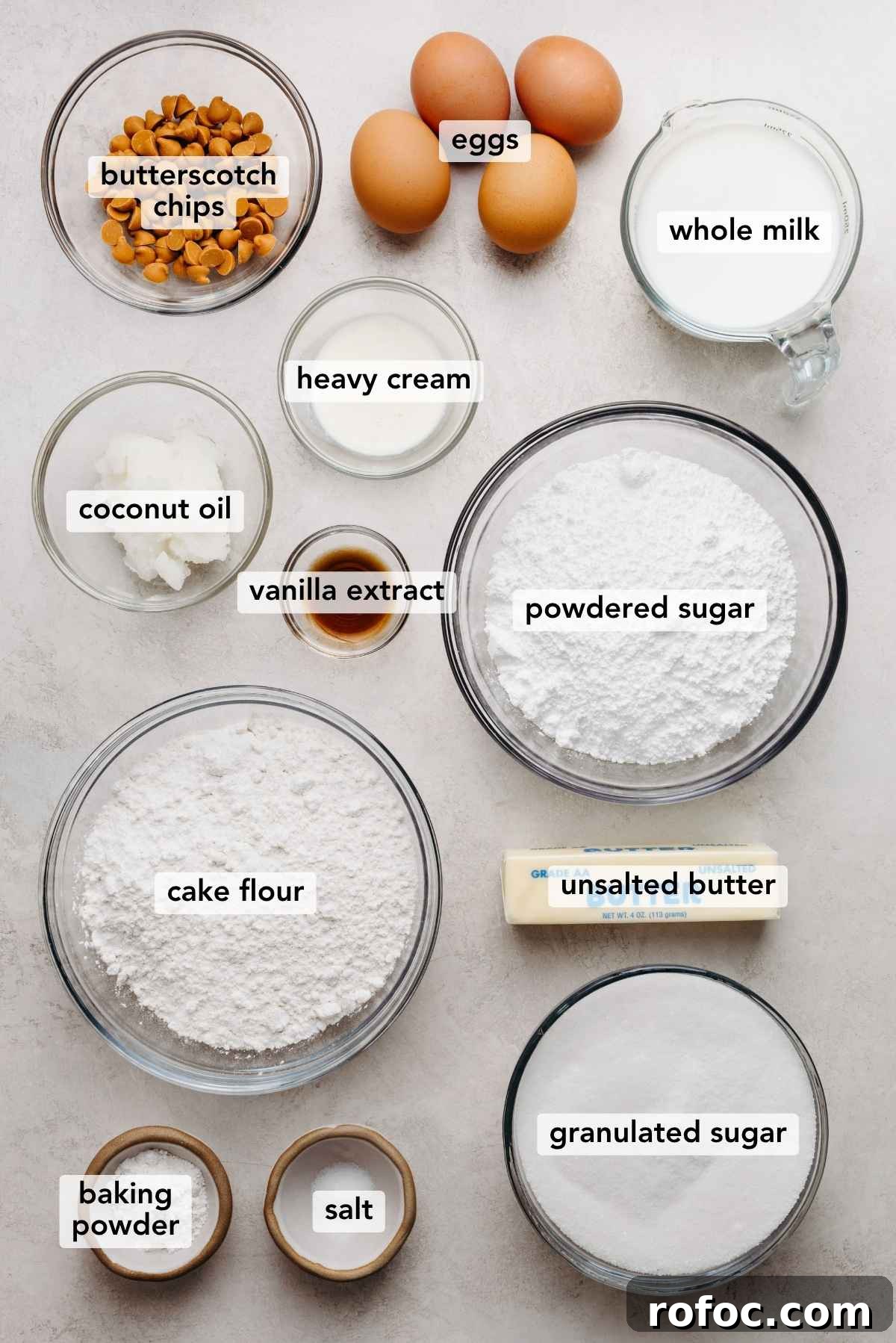 Ingredients in various size bowls labeled as butterscotch chips, eggs, whole milk, coconut oil, heavy cream, vanilla extract, powdered sugar, cake flour, unsalted butter, baking powder, salt, and granulated sugar on a white marbled background.