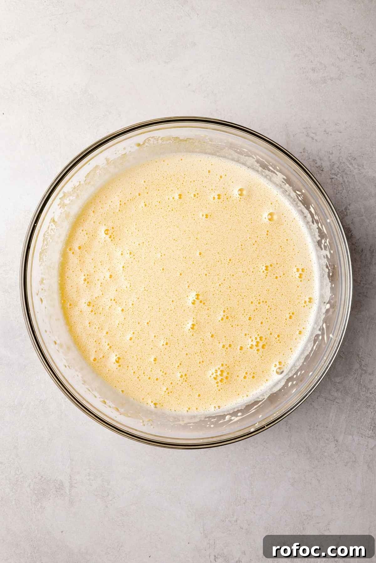 Eggs and granulated sugar whisked together until frothy and doubled in size in a large glass mixing bowl on a white and grey textured background.