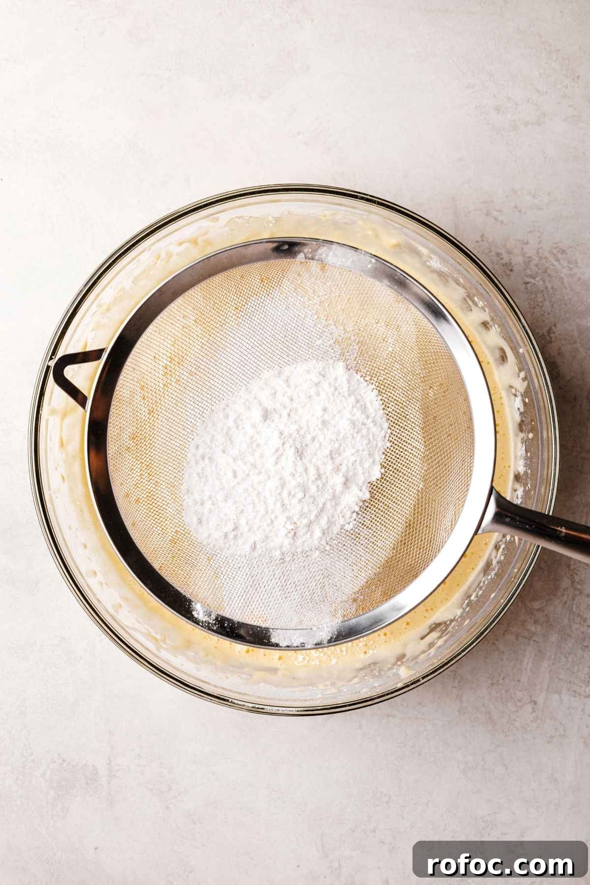 Sifting dry ingredients into a large glass mixing bowl containing sponge cake batter.
