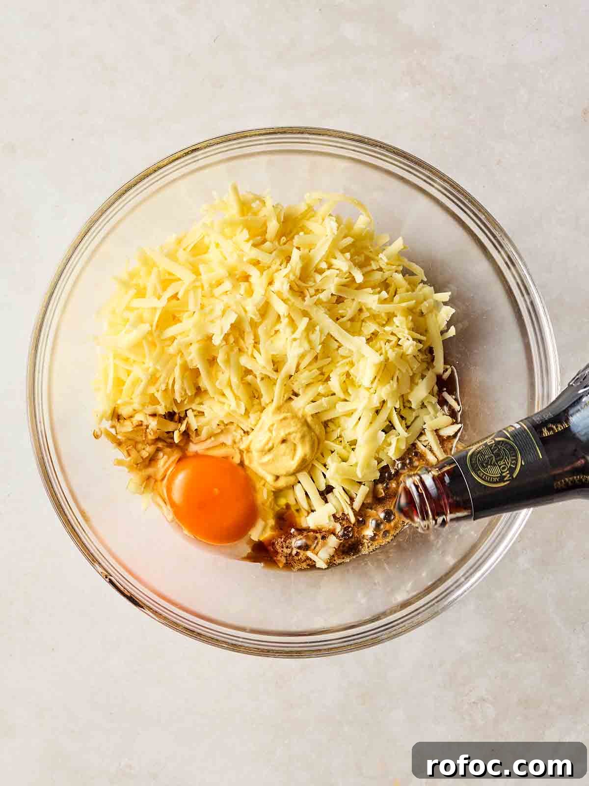 Classic Welsh Rarebit Recipe: Savory Cheese Toast with Ale 5 A glass bowl filled with grated cheese, an eggs and some beer being poured in.