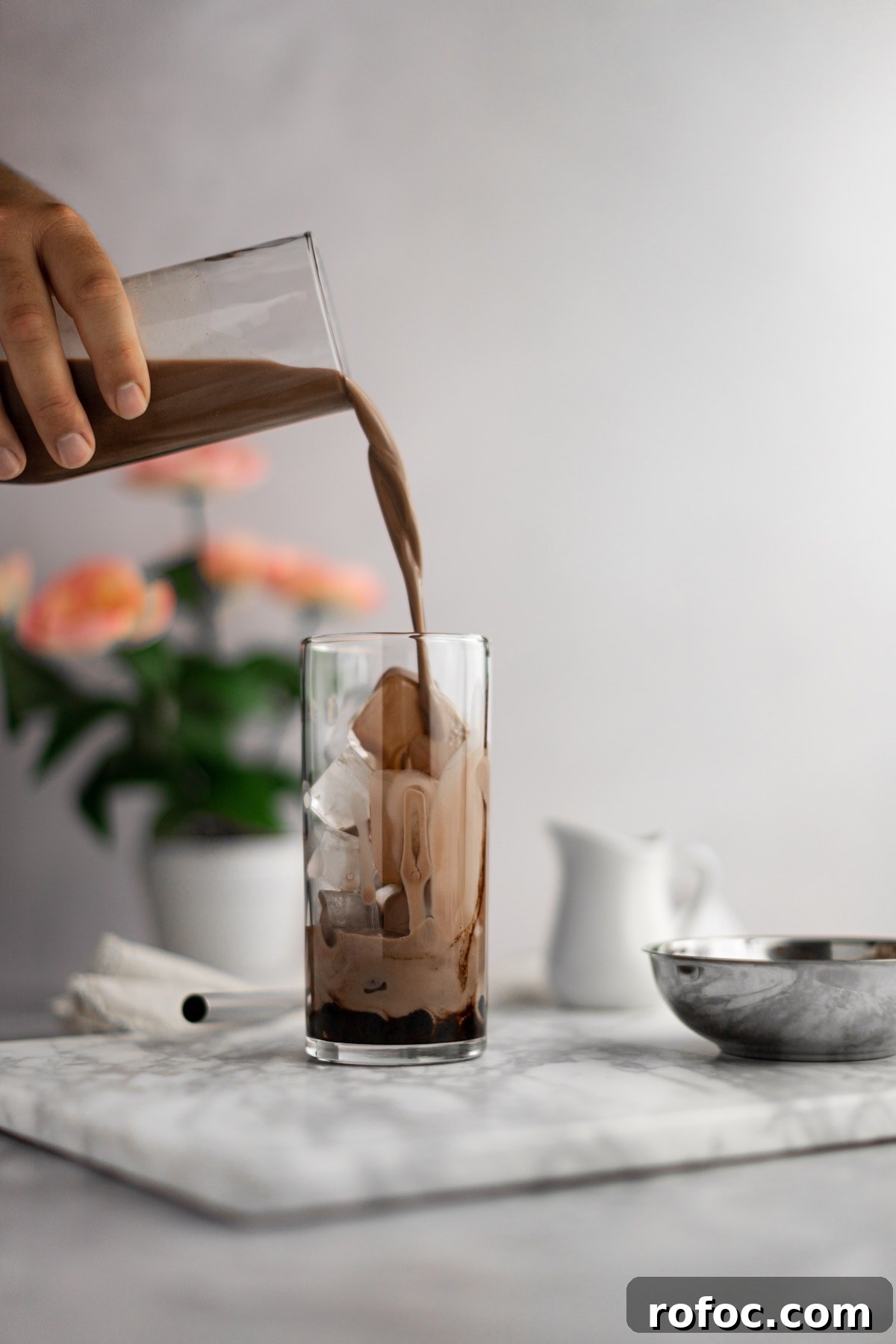 Chocolate Milk Tea Recipe: How to Make Creamy Cocoa Bubble Tea 2 Chocolate milk tea being poured into an empty glass filled with ice and boba, on a white marble board, with pink flowers in the background.