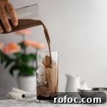 Chocolate milk tea being poured into a glass filled with chocolate soaked boba and ice, pink flowers in the background in front of a white wall.