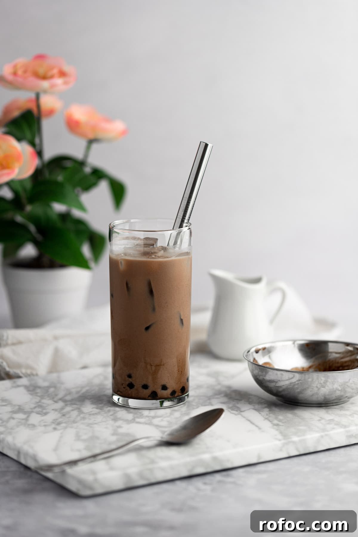 Chocolate Milk Tea Recipe: How to Make Creamy Cocoa Bubble Tea 3 A glass of chocolate milk tea on a grey marble board, next to a metal spoon, with a flower pot in the background.