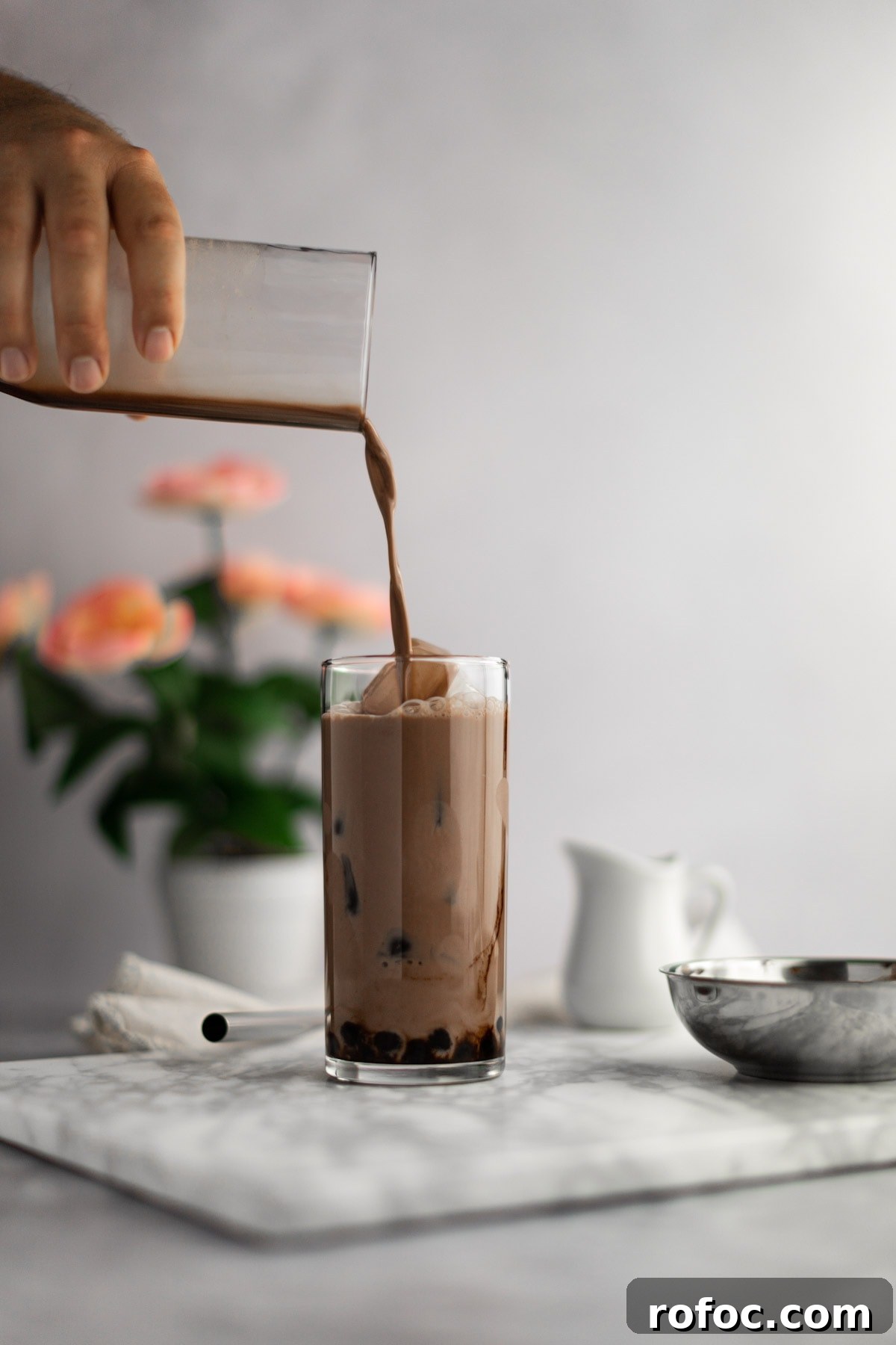 Chocolate Milk Tea Recipe: How to Make Creamy Cocoa Bubble Tea 4 Chocolate bubble tea being poured into an almost full glass, on a grey board, with pink flowers in the background.